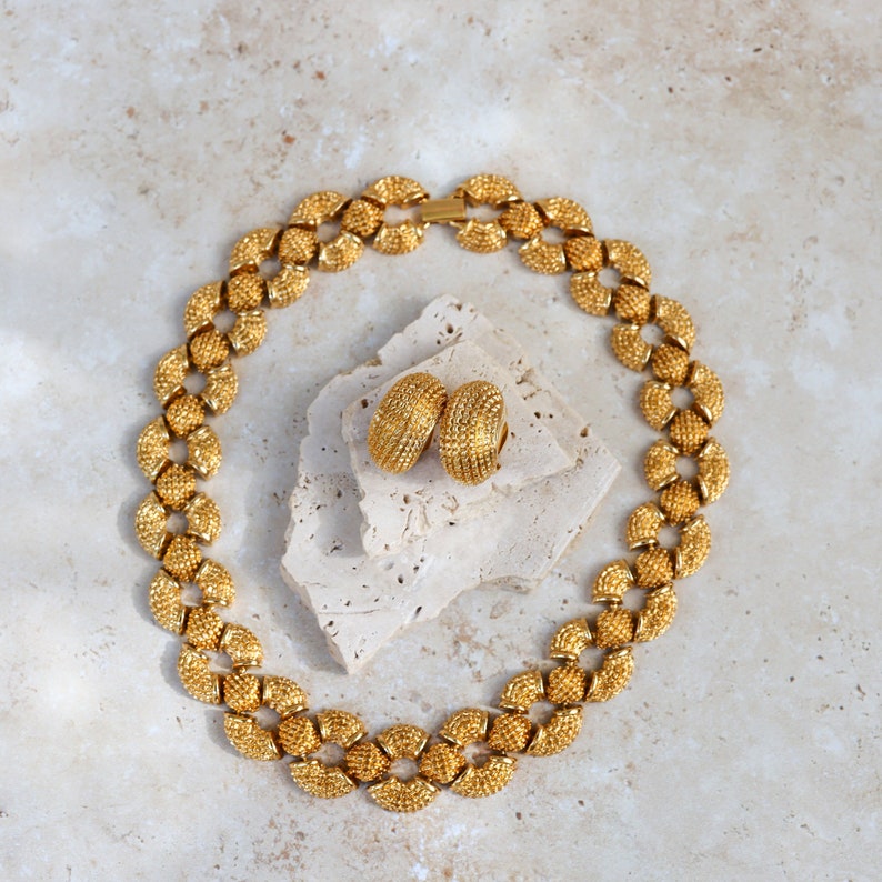 May include: A gold necklace with a chain made of small, round links. The necklace is paired with matching gold earrings. The jewelry is displayed on a light brown surface.
