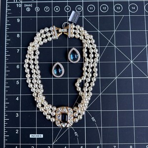 May include: A three-strand pearl necklace with a gold clasp and a square, dark blue and clear stone centerpiece. Matching teardrop-shaped earrings with dark blue stones are also present. The jewelry is displayed on a black grid surface.