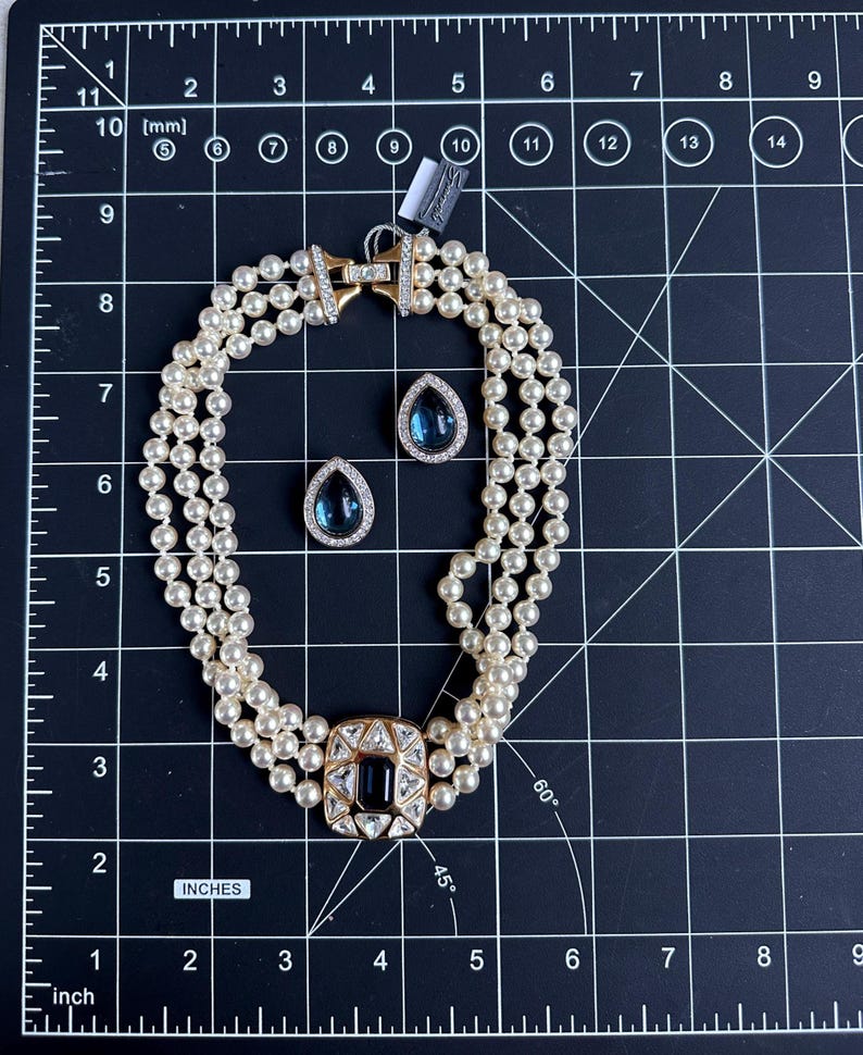 May include: A three-strand pearl necklace with a gold clasp and a square, dark blue and clear stone centerpiece. Matching teardrop-shaped earrings with dark blue stones are also present. The jewelry is displayed on a black grid surface.