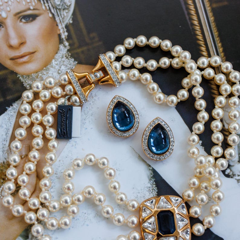 May include: A multi-strand pearl necklace with a gold clasp and matching teardrop earrings with blue gemstones. The earrings and necklace clasp are encrusted with small, clear crystals. A black tag with white text is visible.