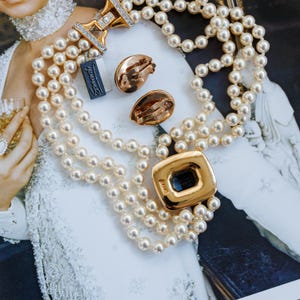 May include: A multi-strand pearl necklace with a gold clasp and a matching gold pendant with a dark blue center. The image also shows a pair of gold clip-on earrings and a tag that says "Stravaganza".