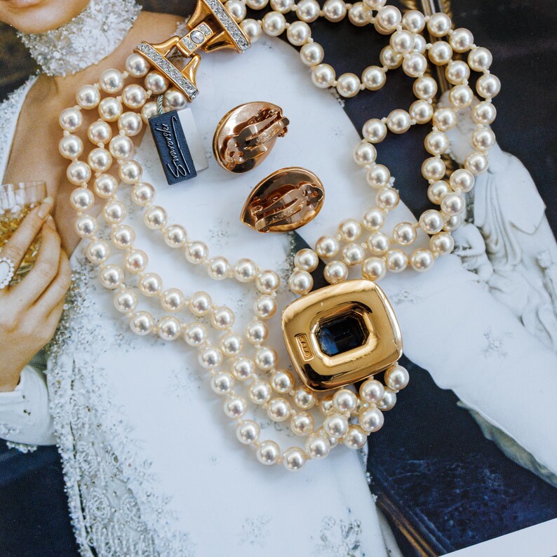 May include: A multi-strand pearl necklace with a gold clasp and a matching gold pendant with a dark blue center. The image also shows a pair of gold clip-on earrings and a tag that says "Stravaganza".