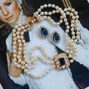May include: A multi-strand pearl necklace with a gold clasp and a square pendant with a dark blue center stone and clear surrounding stones. Matching teardrop-shaped earrings with blue stones and clear accents are also present.