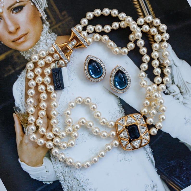 May include: A multi-strand pearl necklace with a gold clasp and a square pendant with a dark blue center stone and clear surrounding stones. Matching teardrop-shaped earrings with blue stones and clear accents are also present.