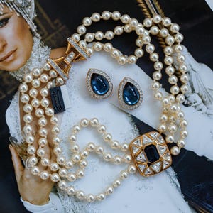 May include: A three-strand pearl necklace with a gold clasp and a large square pendant with a dark blue center stone and clear surrounding stones. Matching teardrop earrings with blue stones and gold settings are also present.