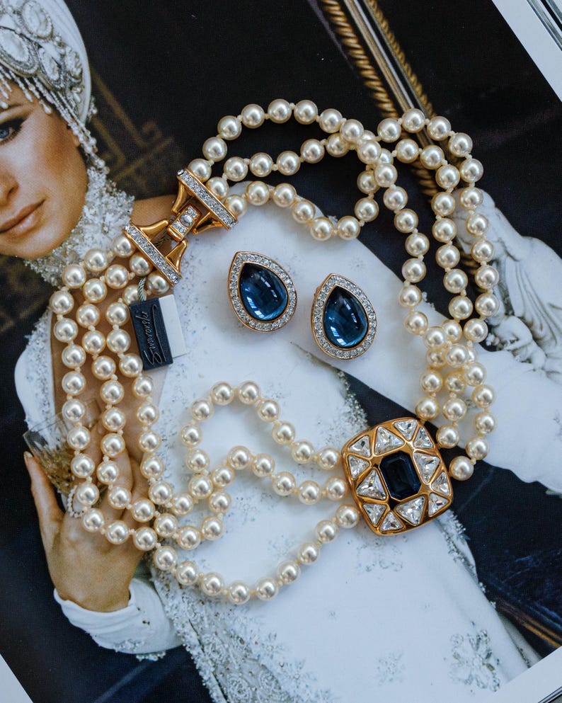 May include: A three-strand pearl necklace with a gold clasp and a large square pendant with a dark blue center stone and clear surrounding stones. Matching teardrop earrings with blue stones and gold settings are also present.