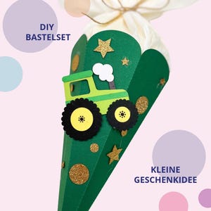 Könnte beinhalten: Ein grüner, kegelförmiger Geschenkkonus mit einem hellgrünen Traktor-Ausschnitt, schwarzen Rädern sowie goldenen Glitzersternen und -kreisen. Die Spitze ist mit weißem Seidenpapier und einer Schnur verschlossen. Text: DIY BASTELSET und KLEINE GESCHENKIDEE.