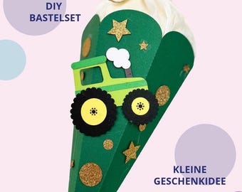 Può includere: Un cono regalo verde con una sagoma di trattore verde chiaro, ruote nere, stelle e cerchi glitterati dorati. La parte superiore è chiusa con carta velina bianca legata con uno spago. Testo: DIY BASTELSET e KLEINE GESCHENKIDEE.
