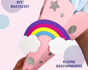 May include: A pink cone-shaped craft project featuring a rainbow, white clouds, and silver glitter stars. The text DIY BASTELSET and KLEINE GESCHENKIDEE is visible. The top of the cone is filled with light blue tissue paper.