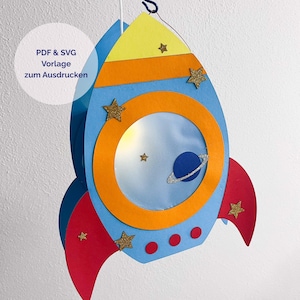 Può includere: Una decorazione a forma di razzo in carta con punta gialla, corpo azzurro e pinne rosse. Include un oblò arancione, un pianeta blu con un anello argentato e stelle glitterate dorate. Testo: PDF & SVG Vorlage zum Ausdrucken.