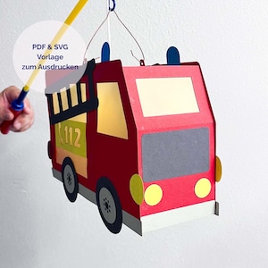 Op de afbeelding: Een rode papieren lantaarn in de vorm van een brandweerwagen met gele ramen, een zwarte ladder en het nummer 112 in het geel. De lantaarn heeft een draadbeugel en een gele stok. De tekst op de afbeelding luidt: PDF & SVG Vorlage zum Ausdrucken.