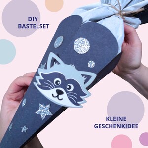 Könnte beinhalten: Ein dunkelblauer, kegelförmiger Geschenkkonus mit einem Waschbären-Ausschnitt, silbernen Glitzersternen und Kreisen sowie einem hellblauen Stoffabschluss, der mit einer Schnur gebunden ist. Der Text DIY BASTELSET und KLEINE GESCHENKIDEE ist abgebildet.