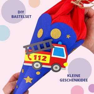 Può includere: Un cono regalo blu con una sagoma di camion dei pompieri rosso dotata di scala e numero 112. La parte superiore è chiusa con carta crespa rossa legata con spago. La superficie blu è decorata con cerchi e stelle glitterate color oro.