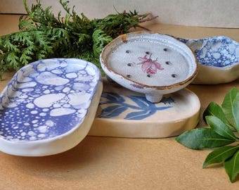 May include: A collection of handmade ceramic soap dishes. One dish features a blue and white bubble pattern, while another has a pink floral design. A third dish has a blue leaf pattern. The dishes are in various shapes and sizes.