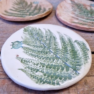 Pode incluir: Um descanso de copo de cerâmica branca com um design de samambaia verde. A samambaia é detalhada com pequenas folhas verdes e um único botão verde.