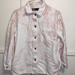 May include: A white and pink tie-dye denim shirt with a button-up front, long sleeves, and a single chest pocket. The shirt has a frayed hem and the label "ZARA" is visible on the collar.