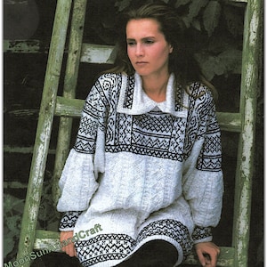 Op de afbeelding: Een wit en zwart patroon gebreide jurk met een brede kraag en lange mouwen. De jurk heeft geometrische ontwerpen rond de kraag, borst en zoom. Het model zit op een verweerde groene houten ladder.