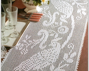 May include: White crocheted table runner with a delicate floral pattern. The runner is laid on a brown surface and features a white vase with pink and white flowers.