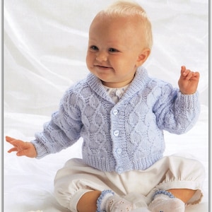 May include: A baby wearing a light blue knitted cardigan with a button fastening and a white knitted hat. The cardigan has a cable knit pattern and the baby is sitting on a white surface.