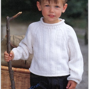 Może przedstawiać: Młody chłopiec w białym swetrze z dzianiny z wzorem warkocza i niebieskich dżinsach. Trzyma patyk w prawej ręce.