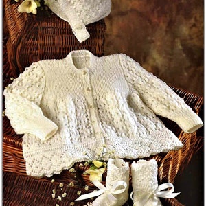 May include: A white knitted baby outfit set including a cardigan, booties, and a bonnet. The set is laid out on a wicker basket with white flowers.