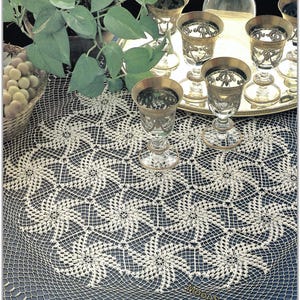 Peut inclure: Une nappe en crochet crème avec un motif floral. La table est dressée avec des verres ornés, un plateau argenté et une bouteille en verre transparent. Un panier de raisins et une plante en pot sont également visibles.