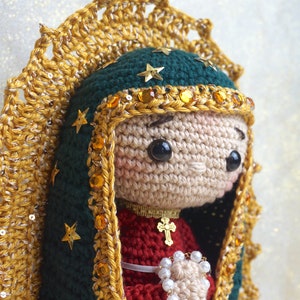 Puede incluir: Mu&ntilde;eca de crochet de la Virgen Mar&iacute;a con una t&uacute;nica verde y dorada con una corona de estrellas. Lleva un rosario blanco en sus manos.
