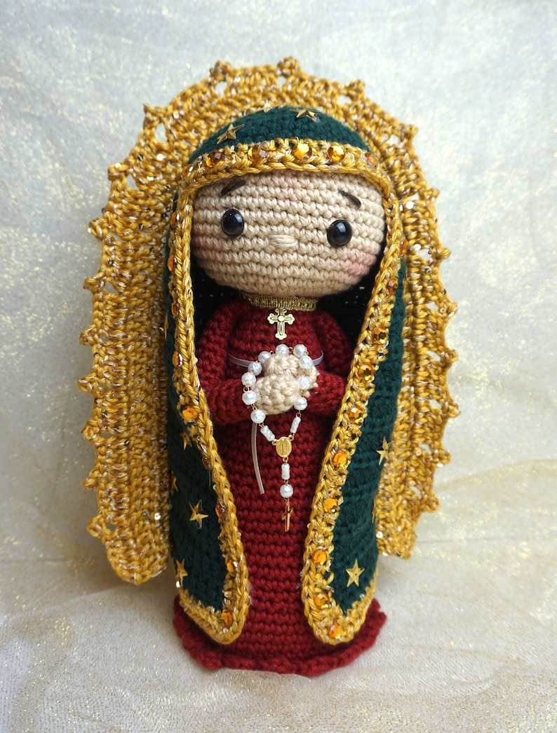 Puede incluir: Mu&ntilde;eca de ganchillo de la Virgen Mar&iacute;a, vestida con un vestido rojo, una capa verde con ribetes dorados y estrellas, y una corona dorada. Lleva un rosario blanco con una cruz dorada.