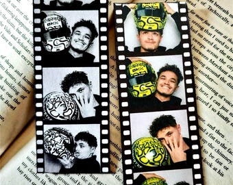 May include: Two vertical film strip bookmarks featuring a series of black and white and color photographs of a person holding a racing helmet. The images capture various poses with the helmet, showcasing detailed patterns and the OKX logo.