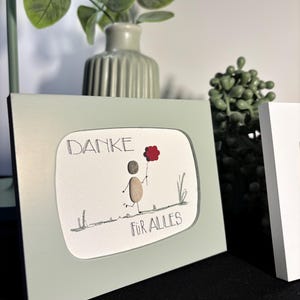 Könnte beinhalten: Ein gerahmtes Kunstwerk mit grünem Hintergrund und weißem Innenraum. Das Kunstwerk zeigt eine einfache Zeichnung einer Person aus Steinen, die eine rote Blume hält. Der Text "DANKE" und "FOR ALLES" ist in Schwarz geschrieben.
