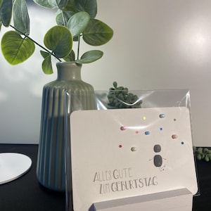 Könnte beinhalten: Eine weiße Karte mit dem Text "Alles Gute zum Geburtstag" und einer kleinen schwarzen Figur mit bunten Punkten drumherum. Die Karte steht auf einem weißen Holzständer.