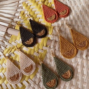 May include: A set of six teardrop-shaped earrings made of small beads. The earrings are in four different colors: orange, black, beige, and green. Each earring has a gold-colored accent at the bottom.