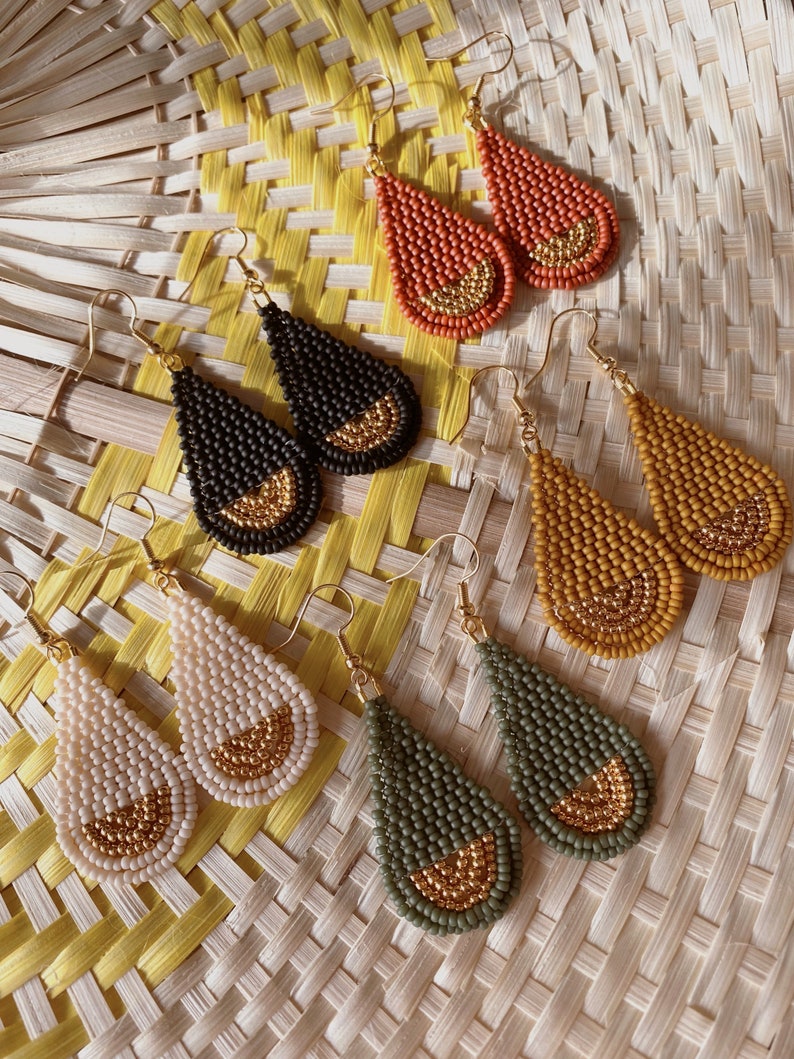 May include: A set of six teardrop-shaped earrings made of small beads. The earrings are in four different colors: orange, black, beige, and green. Each earring has a gold-colored accent at the bottom.