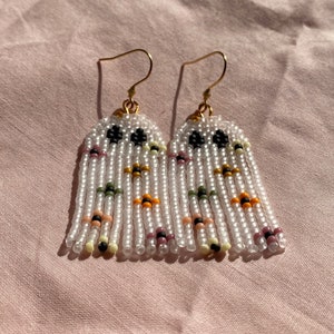 May include: A pair of white beaded earrings with colorful accents. The earrings are made with small, round beads and have a fringe design. The beads are white, black, orange, yellow, green, and pink.