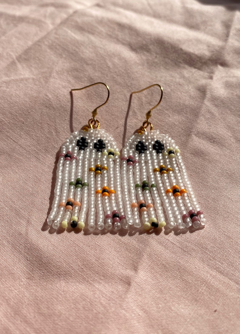 May include: A pair of white beaded earrings with colorful accents. The earrings are made with small, round beads and have a fringe design. The beads are white, black, orange, yellow, green, and pink.
