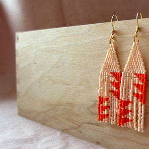 May include: A pair of long, beaded earrings with a peach and orange color scheme. The earrings are hanging from gold hooks.