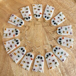 K&ouml;nnte beinhalten: Ein Set mit 10 Paaren wei&szlig;er, geperlter Geisterohrringe mit schwarzen Augen und bunten Akzenten. Die Ohrringe sind in einem Kreis auf einer Holzoberfl&auml;che angeordnet.