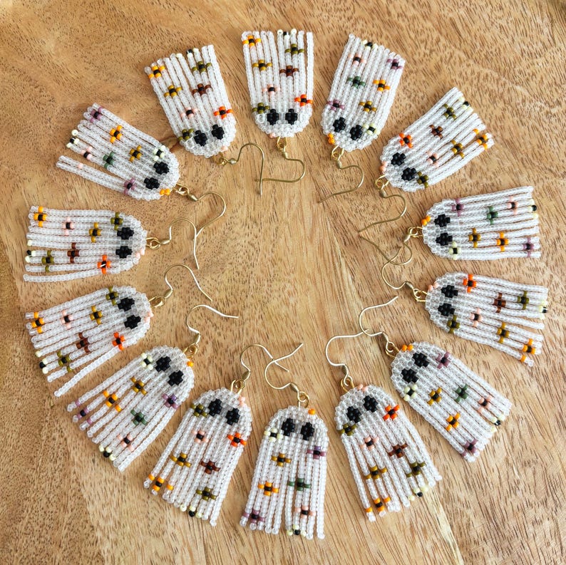 K&ouml;nnte beinhalten: Ein Set mit 10 Paaren wei&szlig;er, geperlter Geisterohrringe mit schwarzen Augen und bunten Akzenten. Die Ohrringe sind in einem Kreis auf einer Holzoberfl&auml;che angeordnet.
