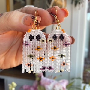 May include: A pair of white beaded ghost earrings with black eyes, orange, purple, and green accents. The earrings are hanging from gold hooks.