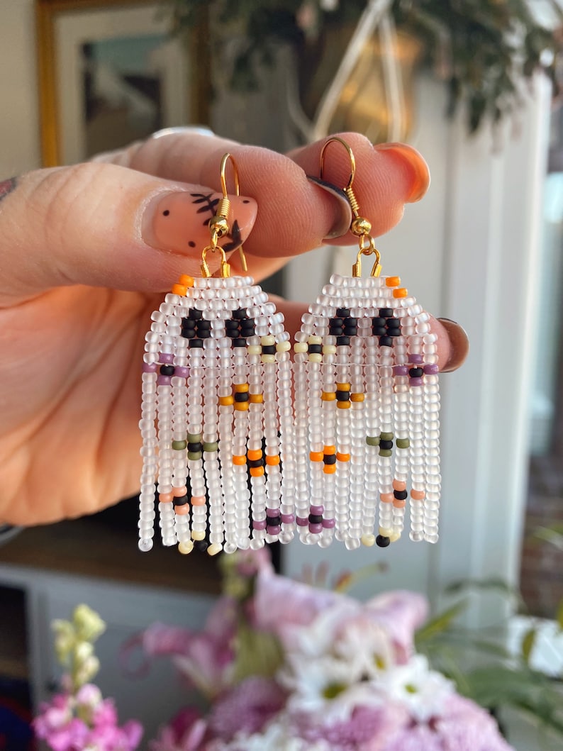 May include: A pair of white beaded ghost earrings with black eyes, orange, purple, and green accents. The earrings are hanging from gold hooks.