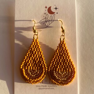 May include: A pair of mustard yellow teardrop-shaped earrings with gold accents. The earrings are made of small beads and have a delicate, bohemian style. The earrings are displayed on a white card with the text "Slow Made Jewellery @palmandluna."