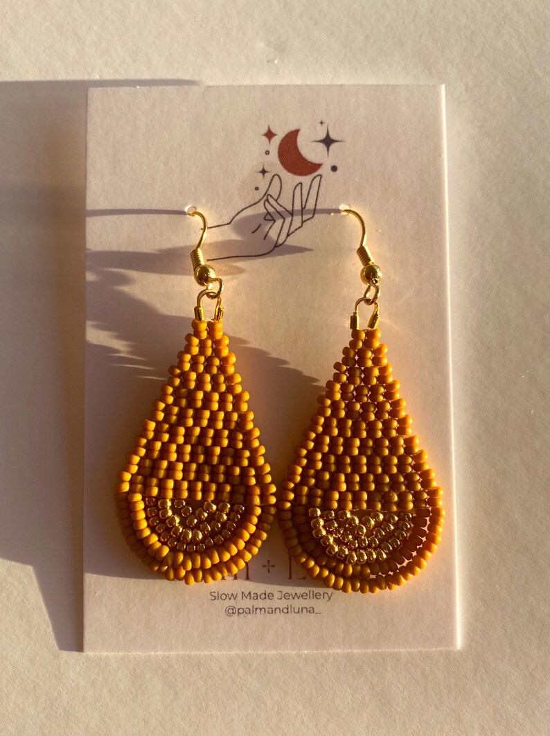 May include: A pair of mustard yellow teardrop-shaped earrings with gold accents. The earrings are made of small beads and have a delicate, bohemian style. The earrings are displayed on a white card with the text "Slow Made Jewellery @palmandluna."