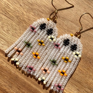 May include: A pair of white beaded earrings with a geometric pattern in black, yellow, pink, and green. The earrings are made with small, round beads and have a fringe-like design.