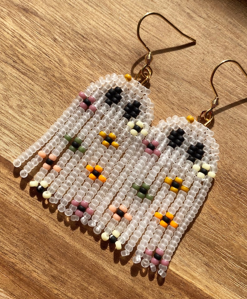 May include: A pair of white beaded earrings with a geometric pattern in black, yellow, pink, and green. The earrings are made with small, round beads and have a fringe-like design.