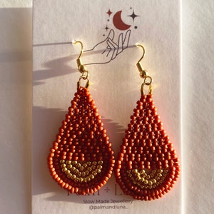 May include: A pair of teardrop-shaped earrings made with orange and gold beads. The earrings have a gold hook closure. The earrings are displayed on a white card with a hand and moon design. The card also includes the text "Slow Made Jewellery @palmandluna_"