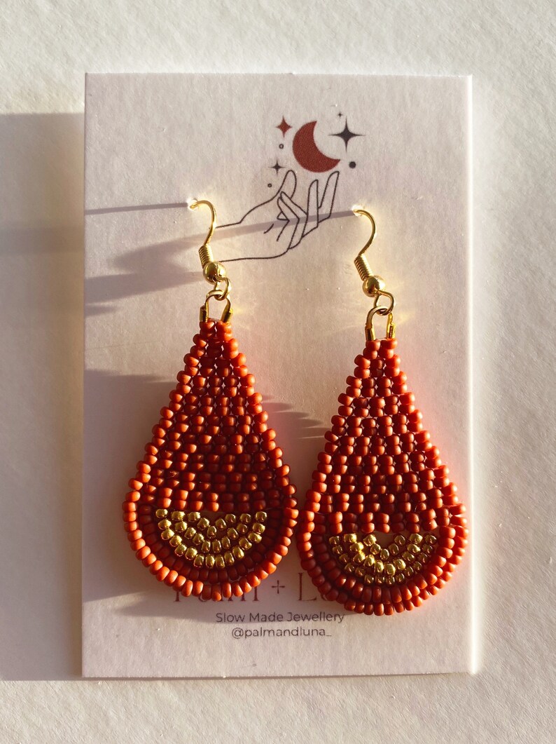 May include: A pair of teardrop-shaped earrings made with orange and gold beads. The earrings have a gold hook closure. The earrings are displayed on a white card with a hand and moon design. The card also includes the text "Slow Made Jewellery @palmandluna_"