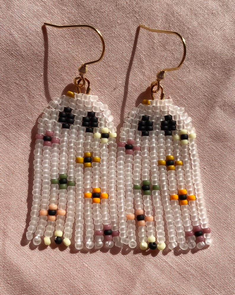 May include: A pair of beaded earrings with a fringe design. The earrings are made with white beads and feature small colorful beads in a floral pattern.