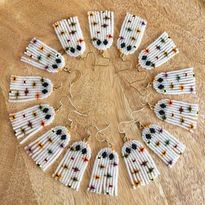 May include: A set of 10 pairs of white beaded ghost earrings with black eyes and colorful accents. The earrings are arranged in a circle on a wooden surface.