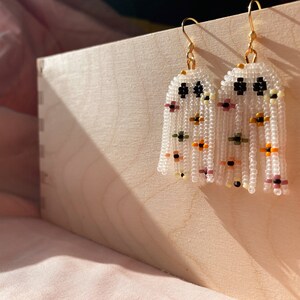 May include: A pair of white beaded earrings with a floral pattern. The earrings are hanging from gold hooks and have a fringe of beads.