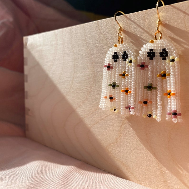 May include: A pair of white beaded earrings with a floral pattern. The earrings are hanging from gold hooks and have a fringe of beads.